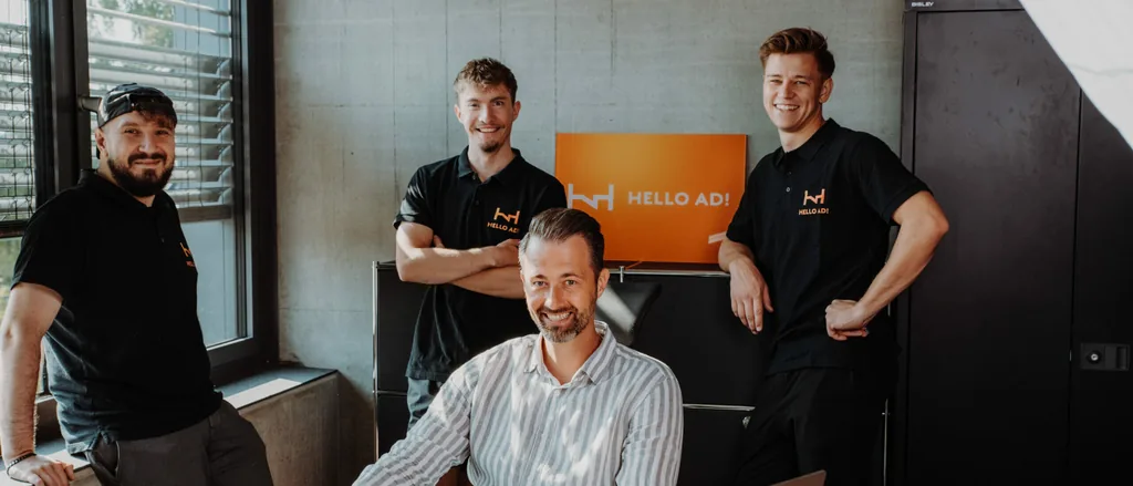 Das Team von HELLO AD! vor der Leuchtschrift im Büro